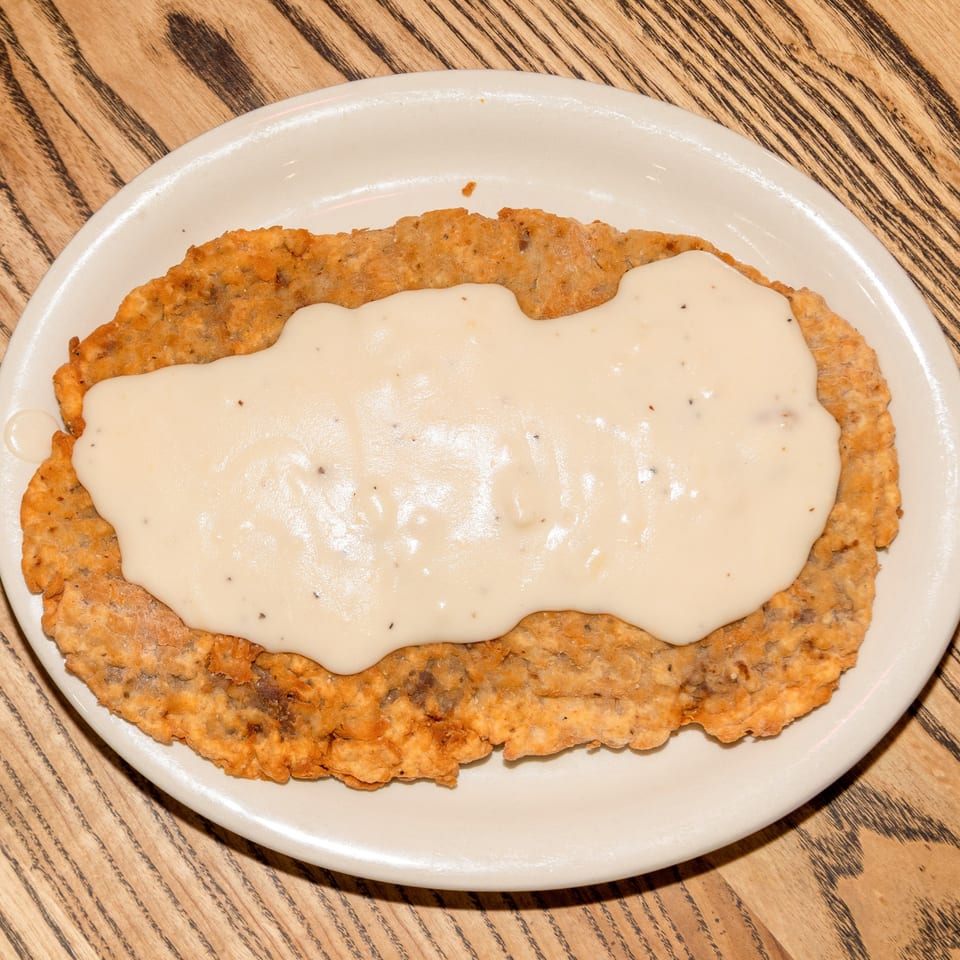 A crispy chicken fried steak covered in creamy white gravy on a white oval plate.