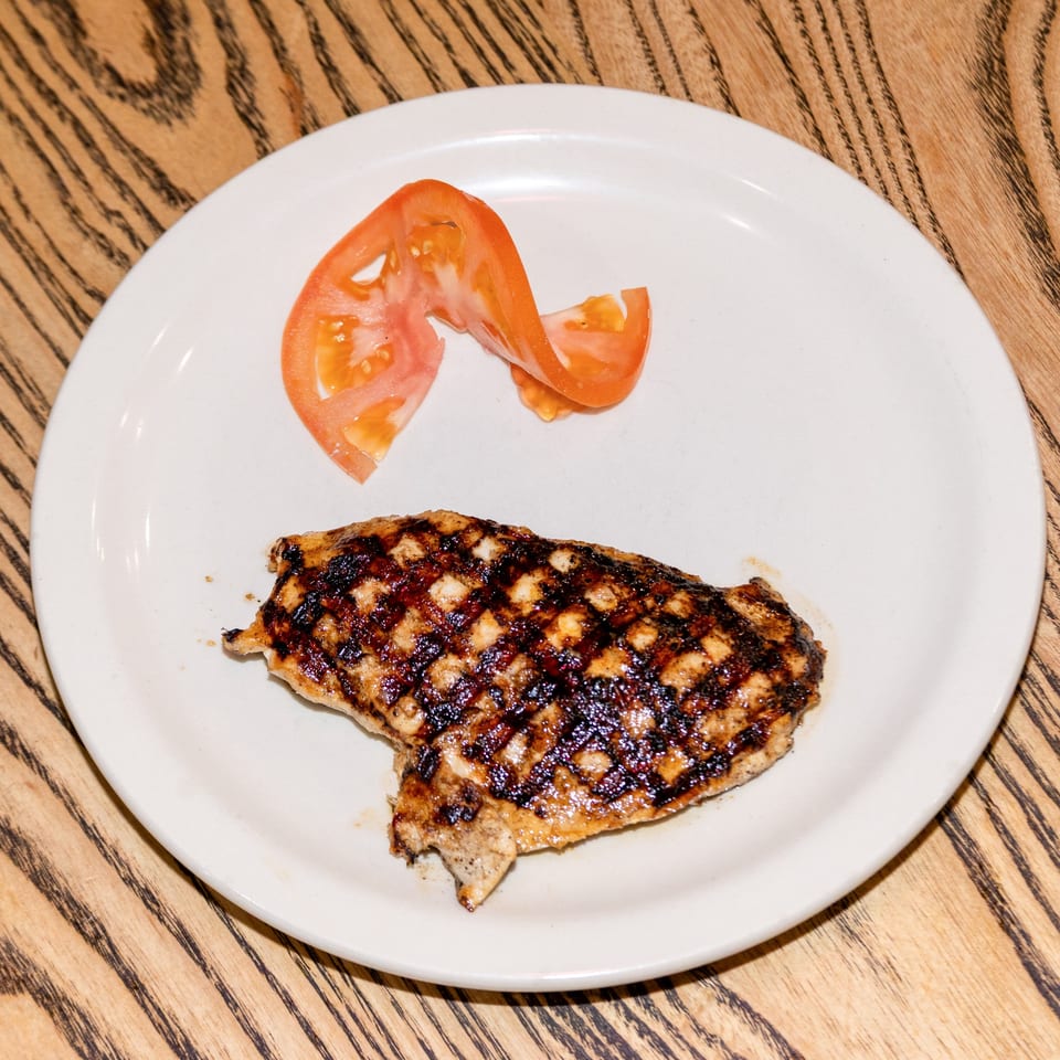 Grilled chicken breast with char marks and a tomato slice on a white plate at Houligan's Corner.