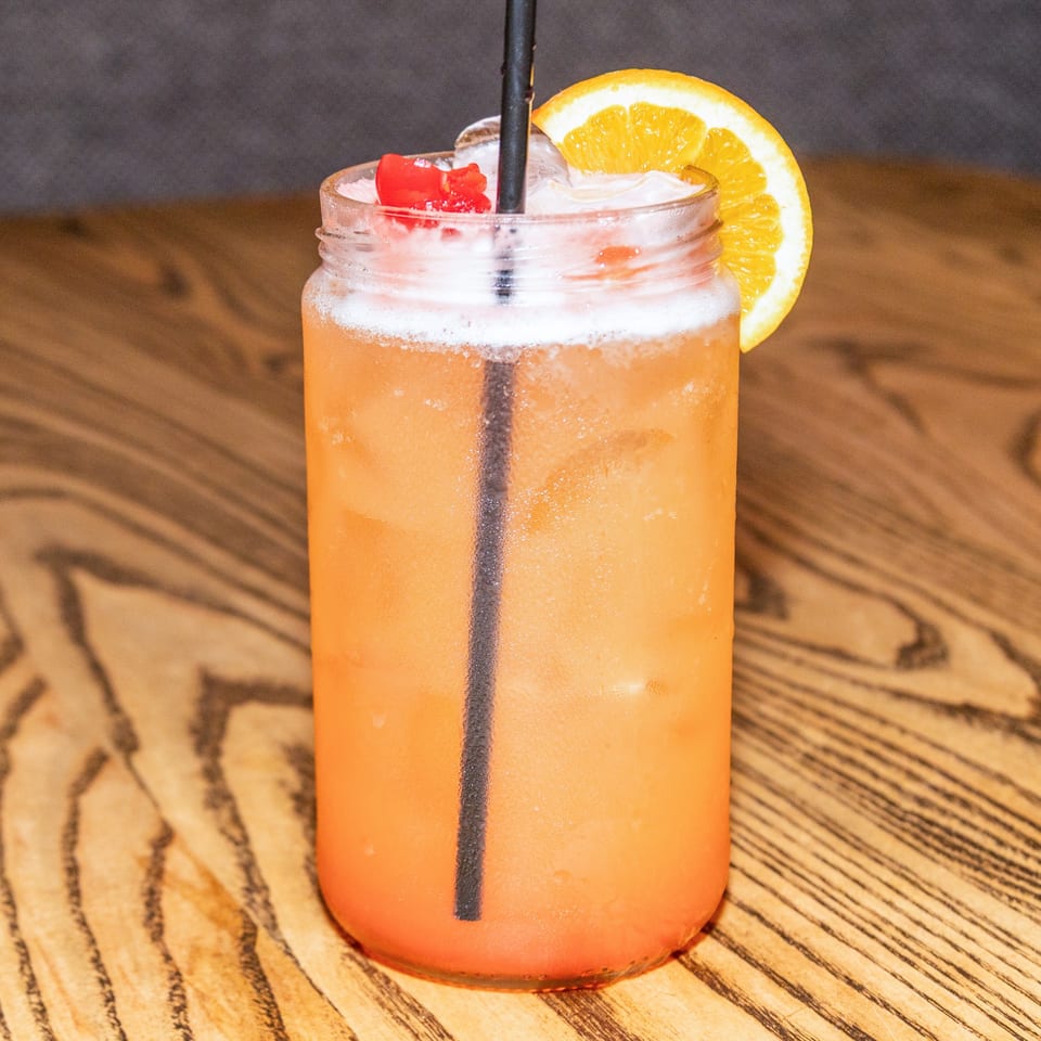 A refreshing Houligan's Corner cocktail in a mason jar, garnished with an orange slice and cherries.