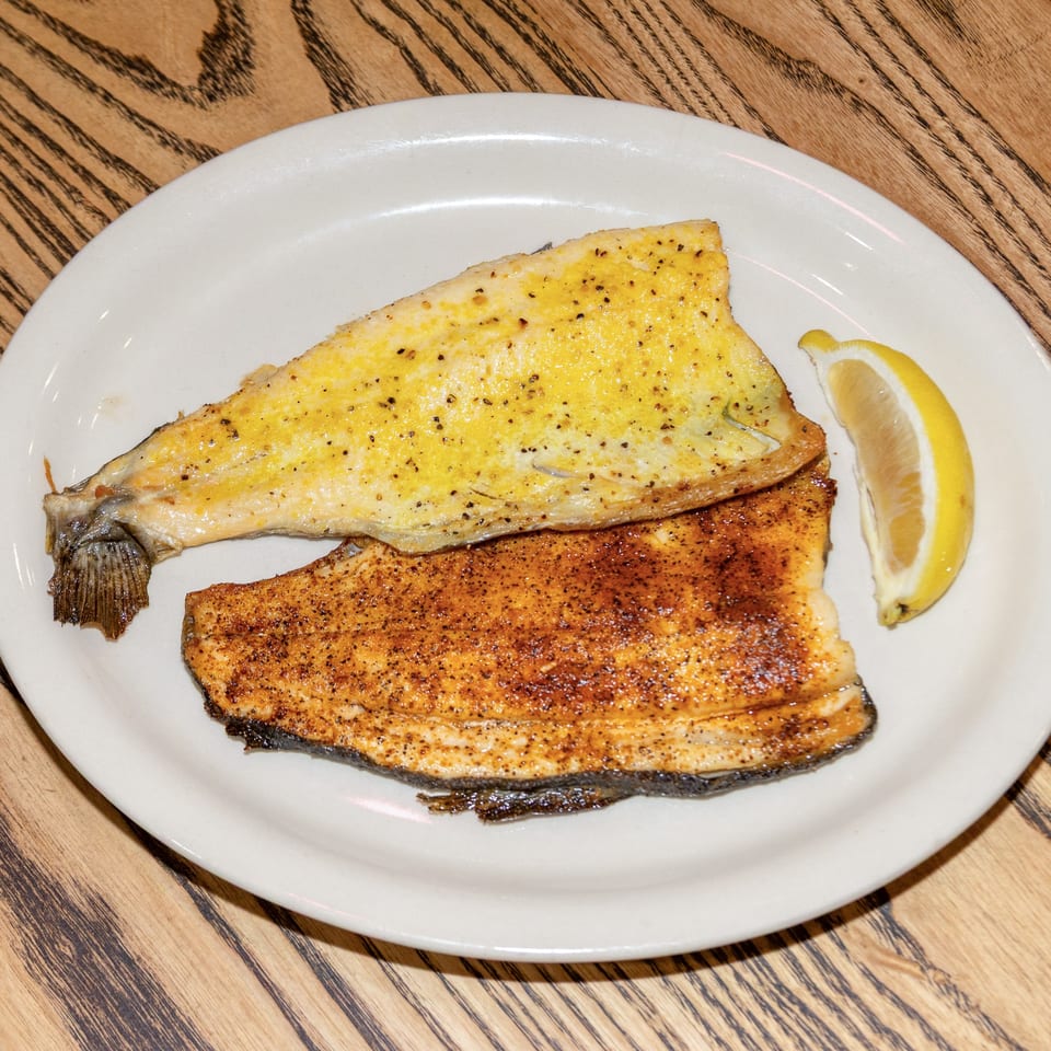 Two fillets of fish, one buttered and one blackened, with a lemon wedge on a white plate.