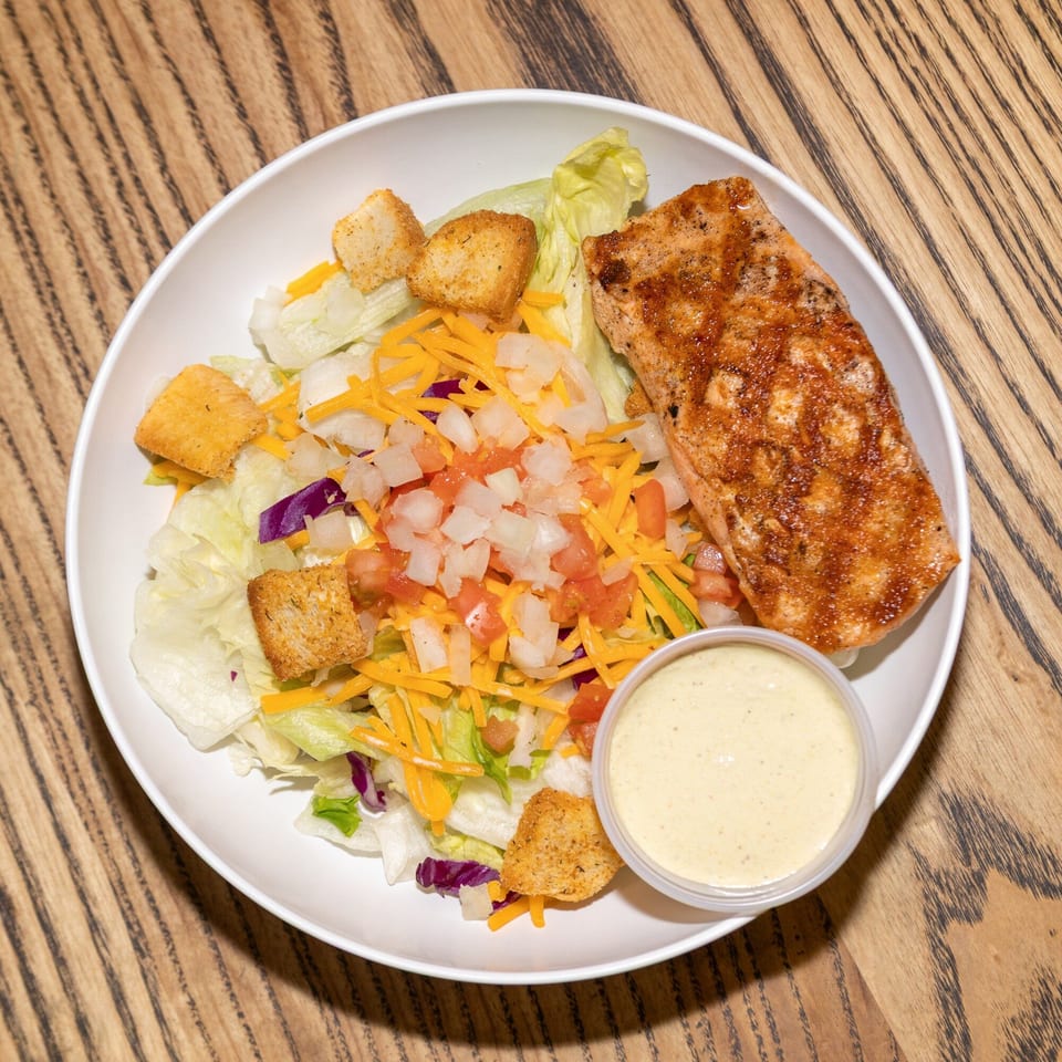 A Houligan's Corner salmon salad with lettuce, cheese, tomatoes, croutons, and a side of dressing.