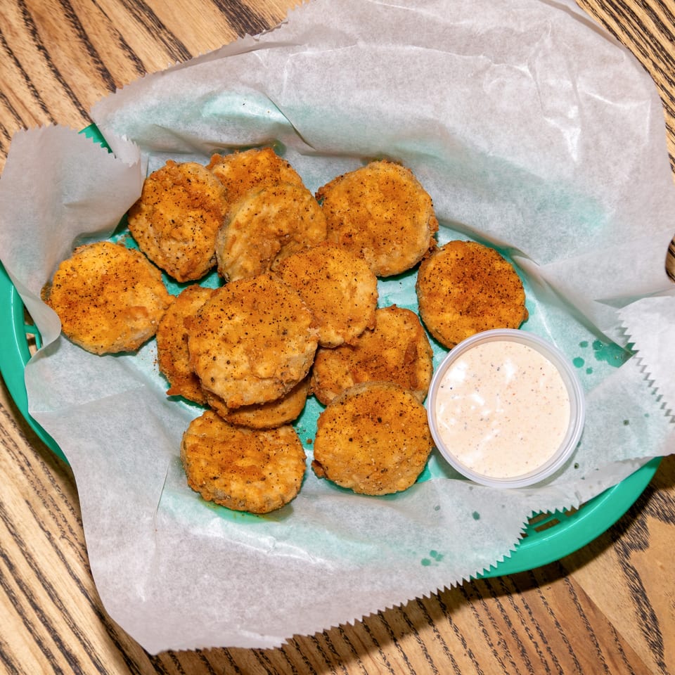 A basket of Houligan's Corner fried pickles with a side of dipping sauce on a wooden table.
