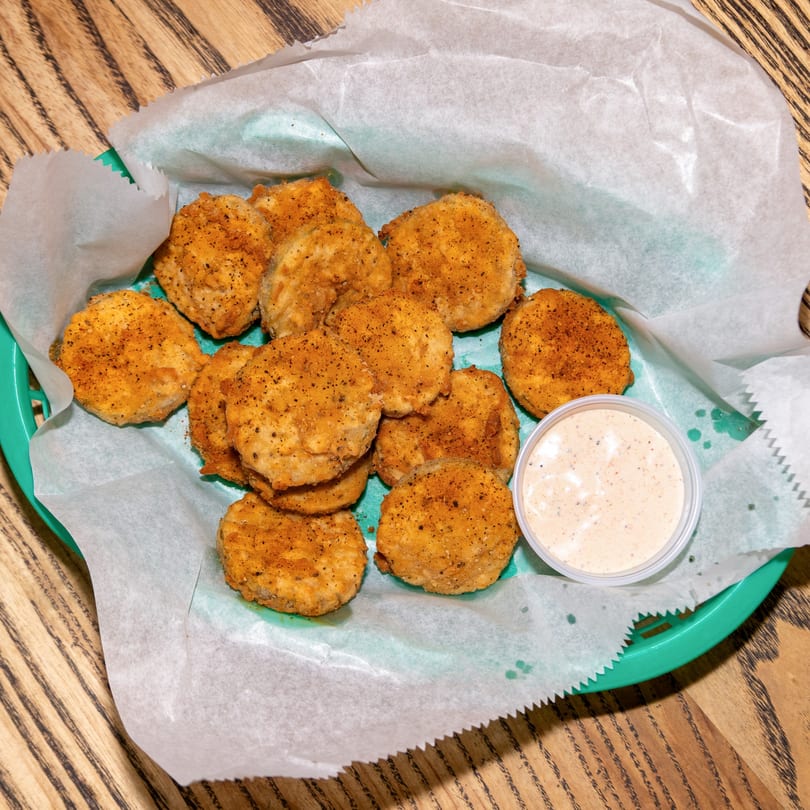 A basket of Houligan's Corner fried pickles with a side of dipping sauce on a wooden table.