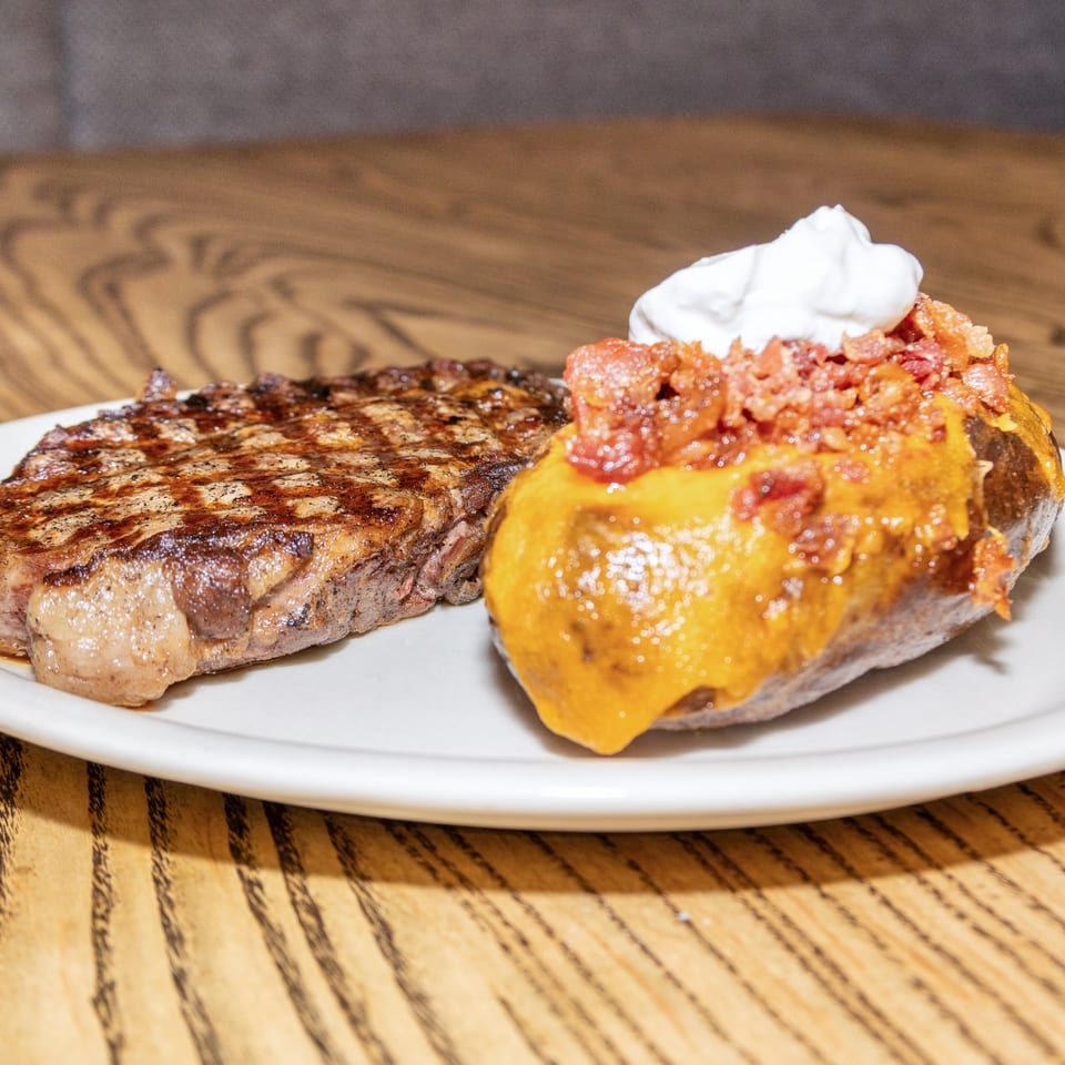 Grilled steak and a loaded baked potato with cheese, bacon, and sour cream on a white plate.